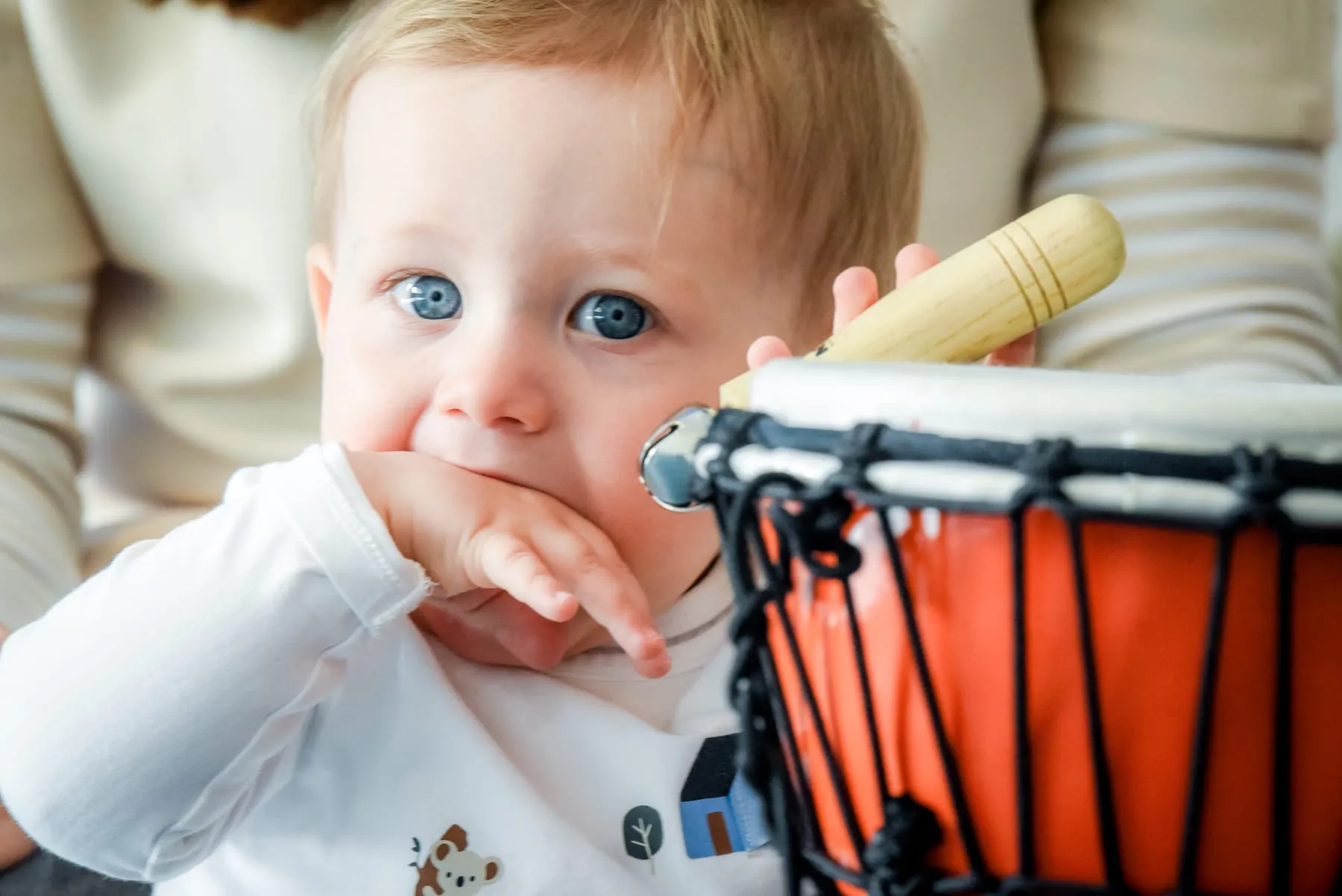 Ein Baby mit der Hand am Mund schaut in die Kamera, daneben eine orangefarbene Djembe mit einem hellen Holzschlägel