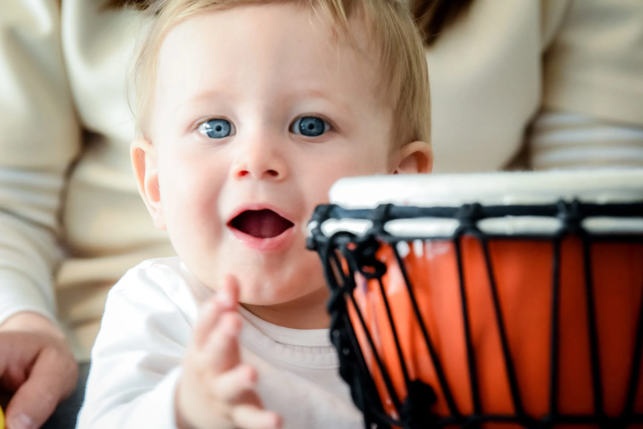 Ein Baby klatscht in die Hände und schaut mit offenem Mund, vor ihm eine orangefarbene Trommel, hinter ihm sitzt ein Erwachsener