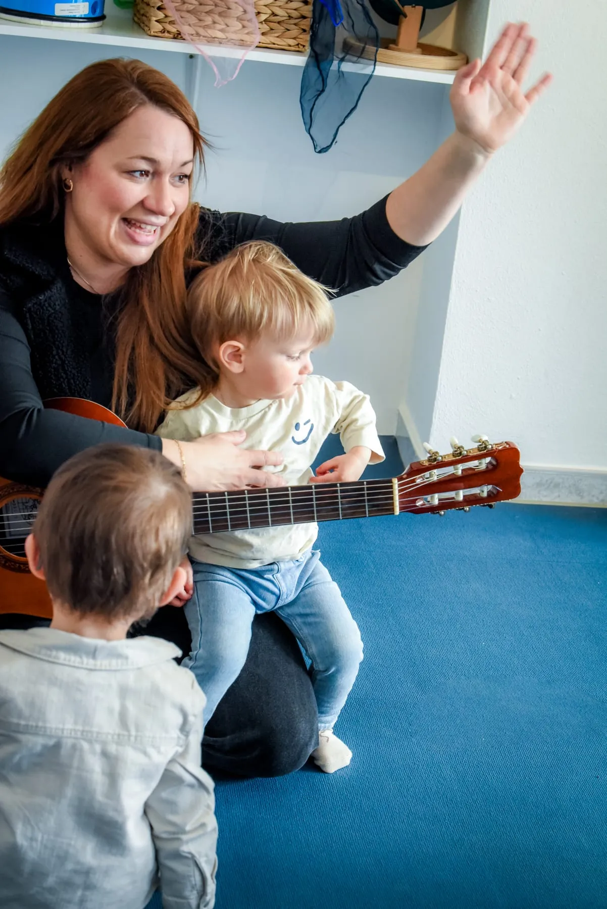 Eine Frau mit Gitarre hebt die Hand, auf ihrem Schoß sitzt ein Kleinkind, vor ihr steht ein weiteres Kind mit dem Rücken zur Kamera