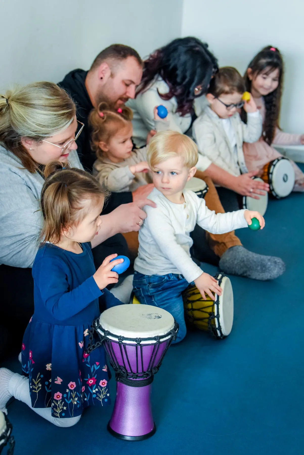 Kinder und Erwachsene sitzen eng nebeneinander auf dem Boden und spielen auf Djemben und mit Rassel-Eiern, vorn ein Mädchen an einer lila Djembe