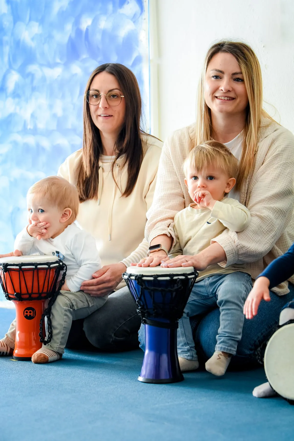 Zwei Frauen sitzen nebeneinander auf dem Boden, vor ihnen je ein Baby an einer Djembe, die rechte Frau hält ihr Kind im Arm