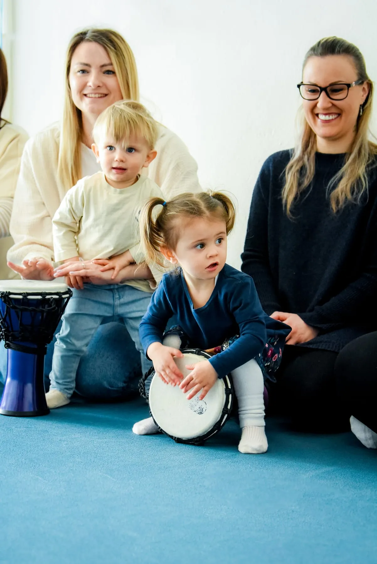 Ein Mädchen hockt über einer kleinen Trommel und legt beide Hände aufs Fell, dahinter sitzen zwei lächelnde Frauen, eine hält ein Kleinkind