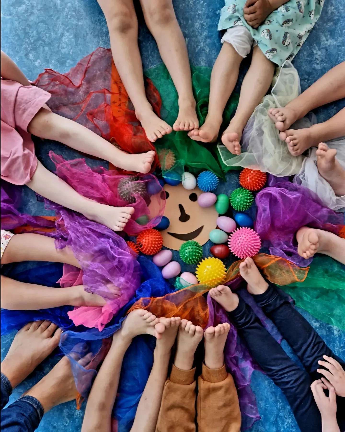 Nackte Füße im Kreis um bunte Tücher, Igelbälle und Rassel-Eier, in der Mitte ein lachendes Gesicht aus Pappe, von oben fotografiert