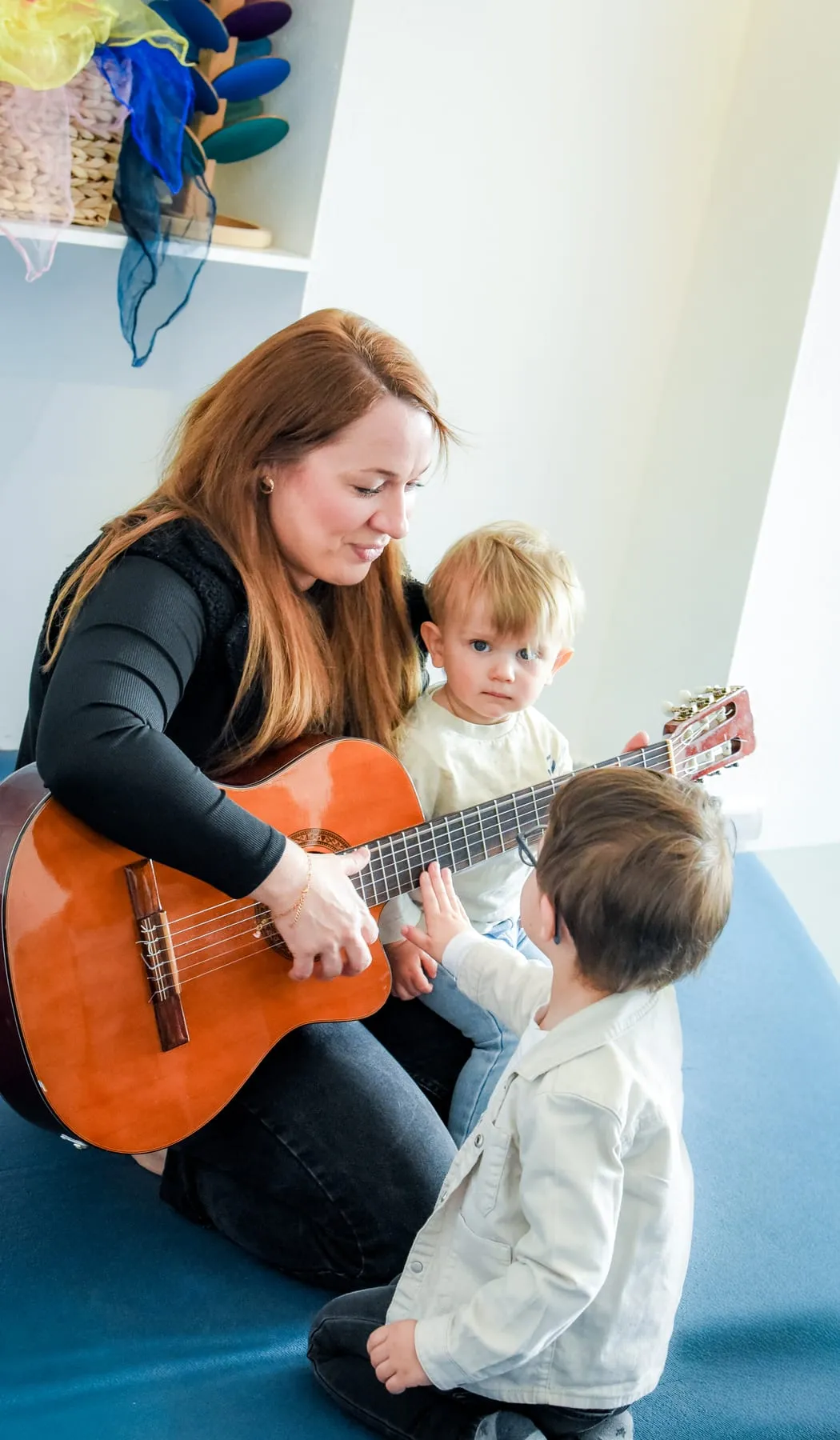 Eine Frau spielt Gitarre, ein Kleinkind legt die Hand auf die Saiten, ein zweites sitzt dicht neben ihr