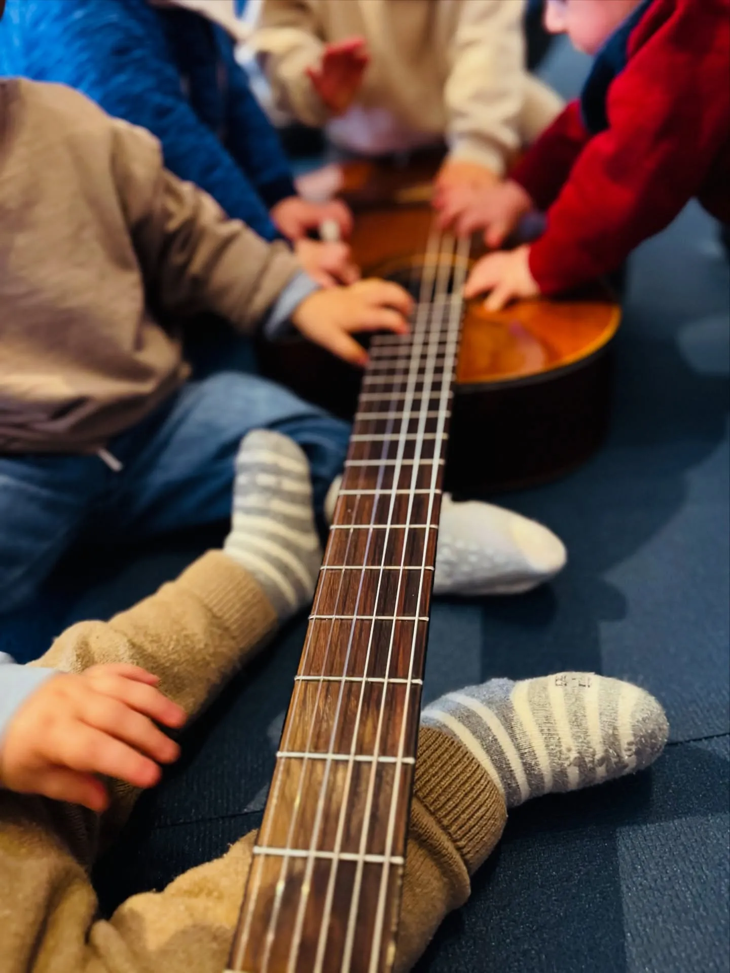 Eine Gitarre liegt auf dem Boden, mehrere Kleinkinder sitzen darum und legen die Hände auf Saiten und Korpus