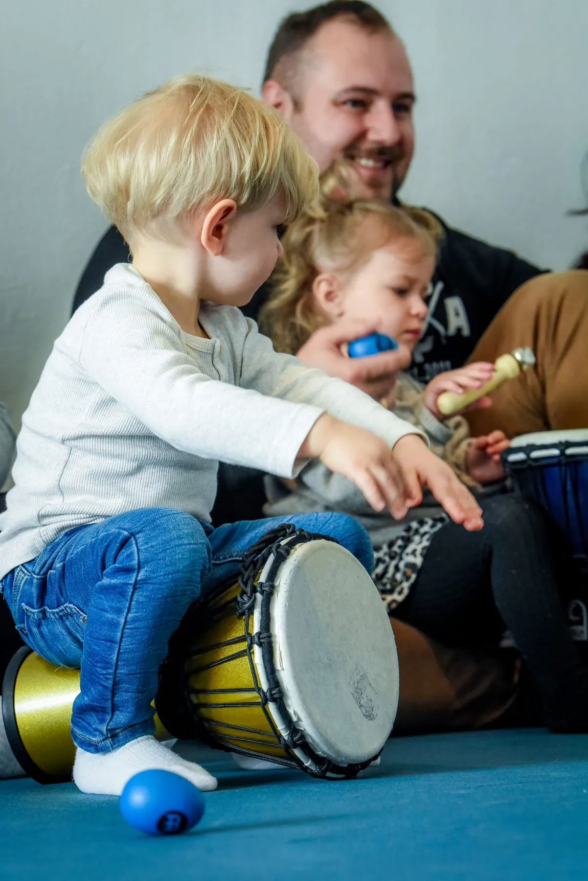 Ein Junge sitzt auf einer Djembe und schlägt mit beiden Händen darauf, hinter ihm lacht ein Mann, daneben sitzt ein Mädchen mit einem blauen Rassel-Ei