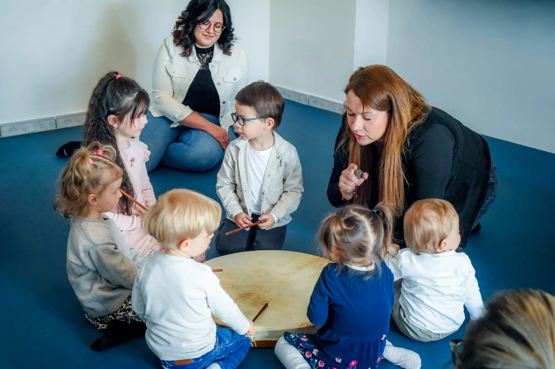 Zwei Frauen sitzen mit mehreren Kleinkindern im Kreis um eine große Rahmentrommel, eine der Frauen spricht zu den Kindern