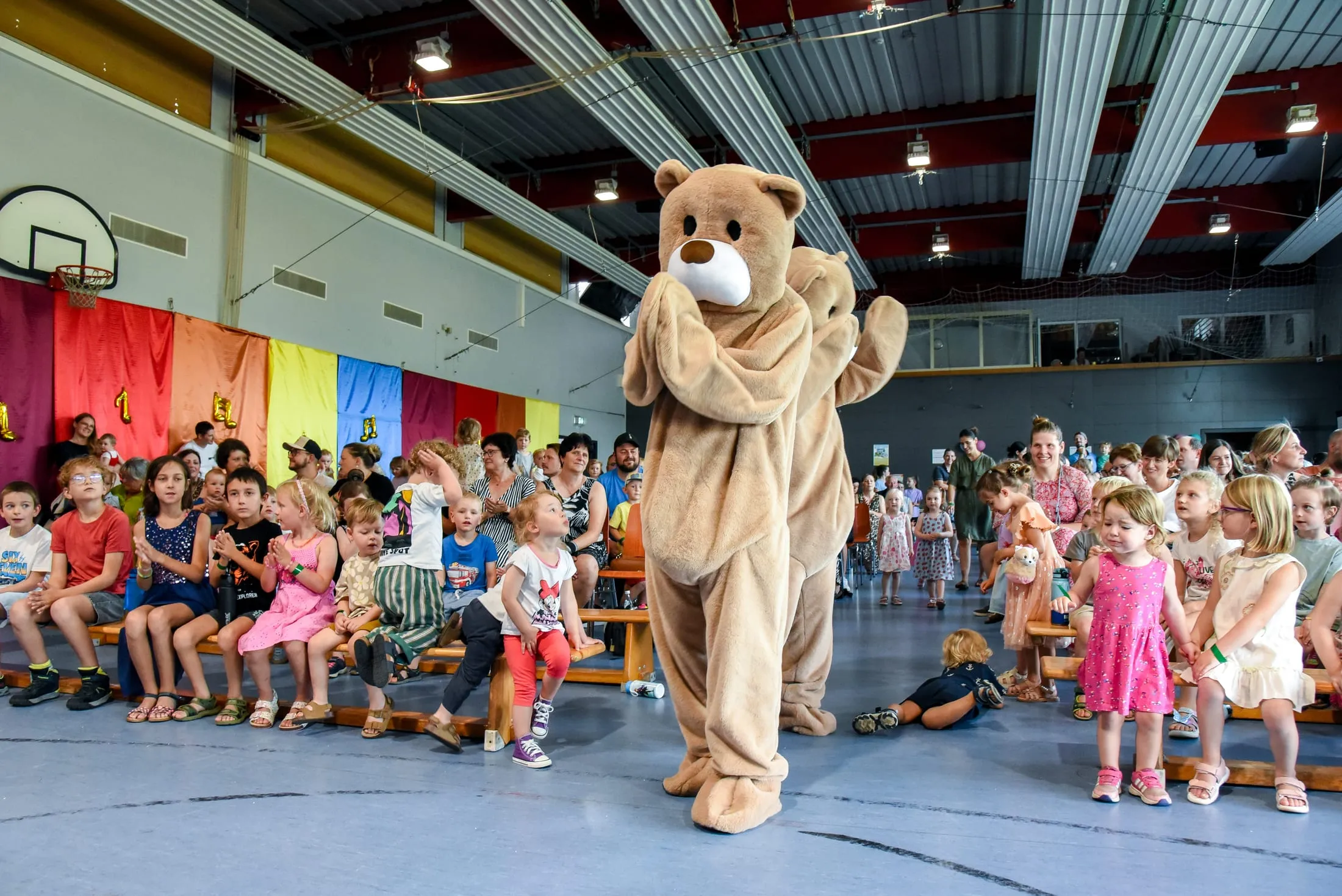 Zwei Bären-Maskottchen gehen durch eine Halle voller Kinder und Erwachsener auf Bänken, einige Kinder klatschen