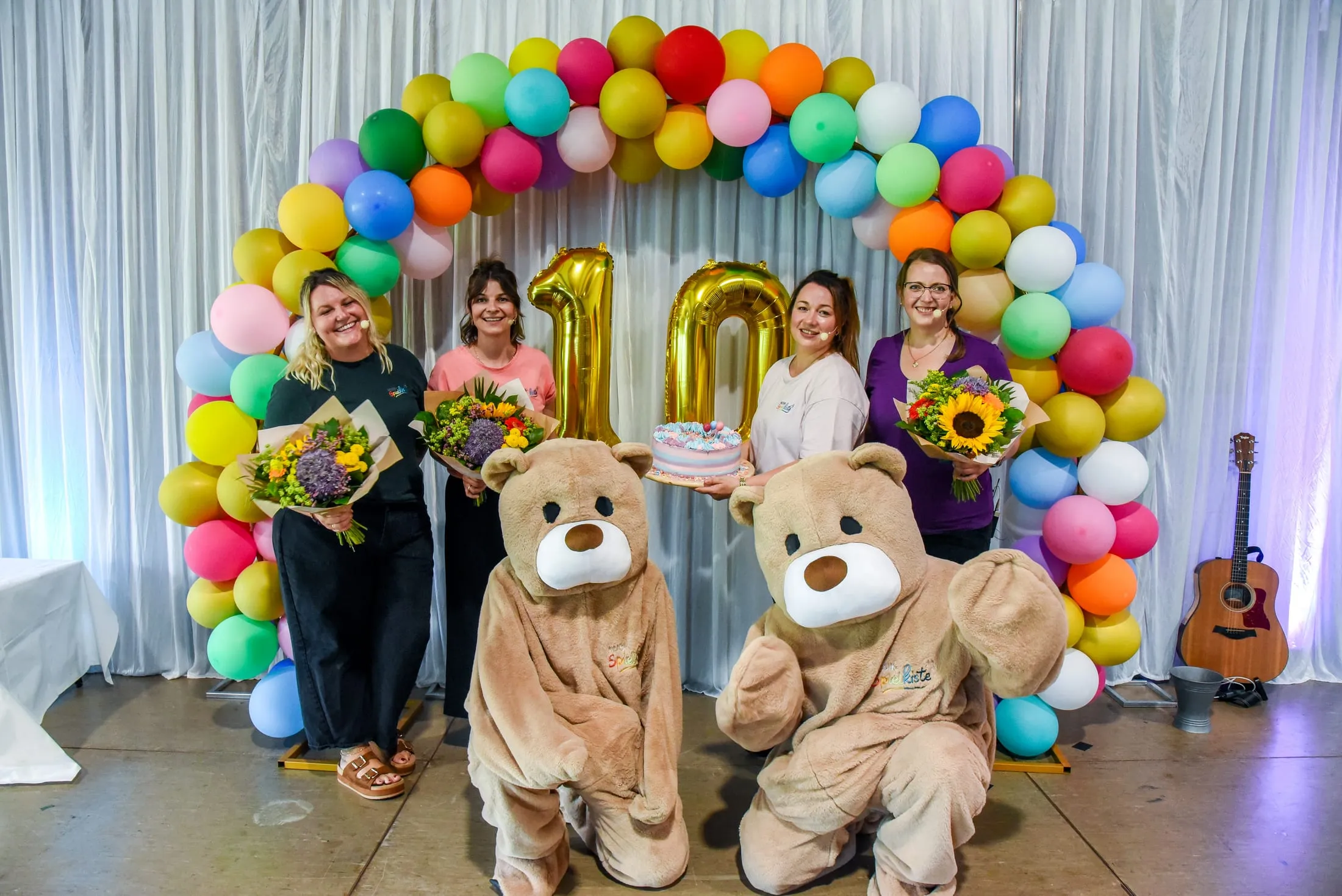 Vier Frauen vom Team mit drei Blumensträußen und einer Torte unter einem bunten Ballonbogen mit einer goldenen 10, davor knien zwei Bären-Maskottchen