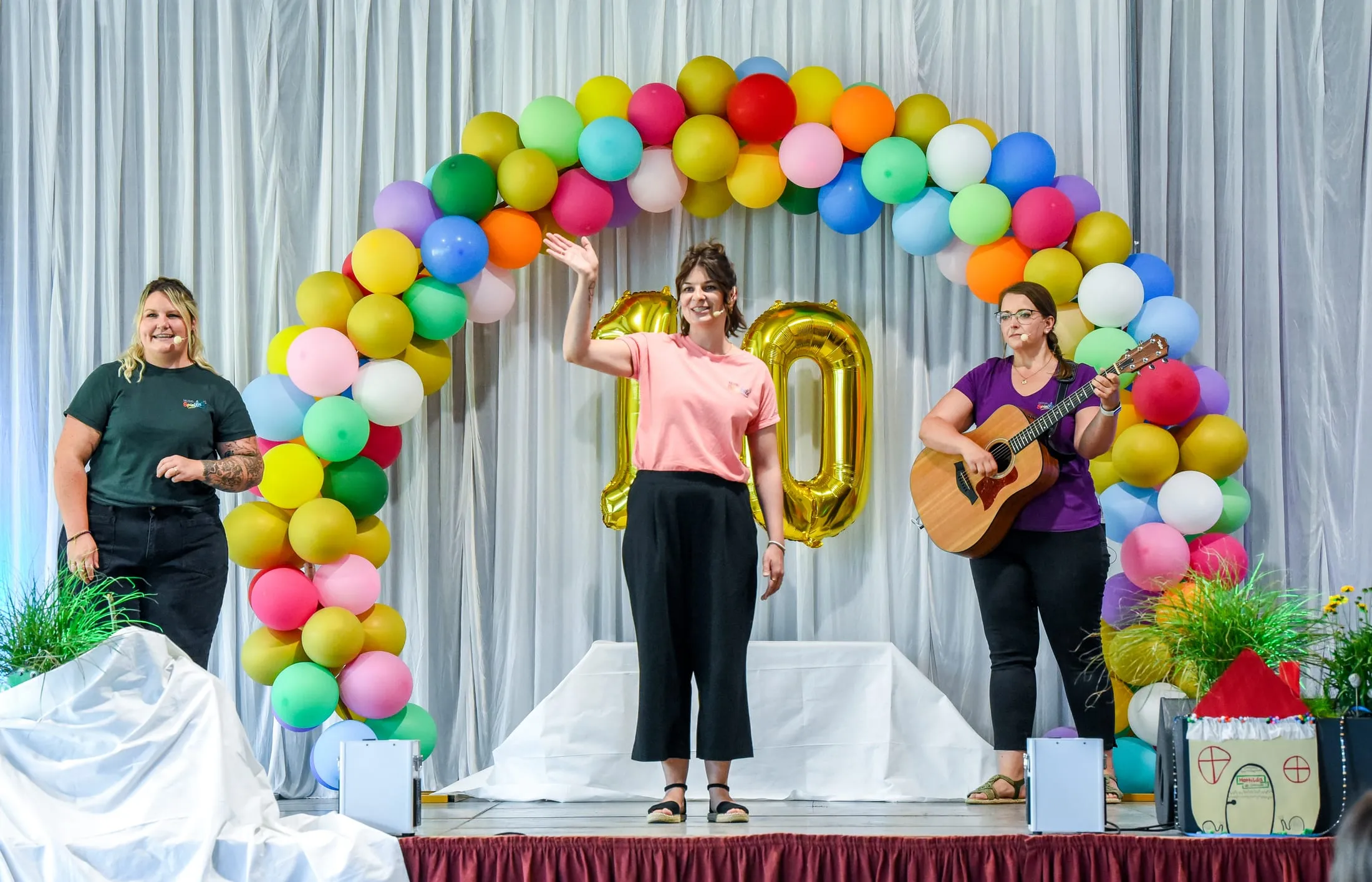 Drei Frauen vom Team auf der Bühne unter einem bunten Ballonbogen mit einer goldenen 10, eine winkt, eine spielt Gitarre