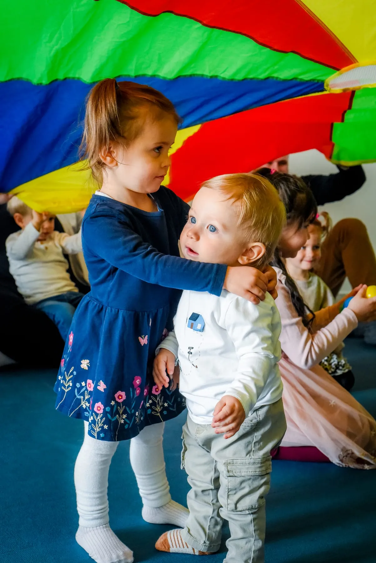 Ein Mädchen in dunkelblauem Kleid legt die Arme um einen kleineren Jungen, über ihnen ein buntes Schwungtuch