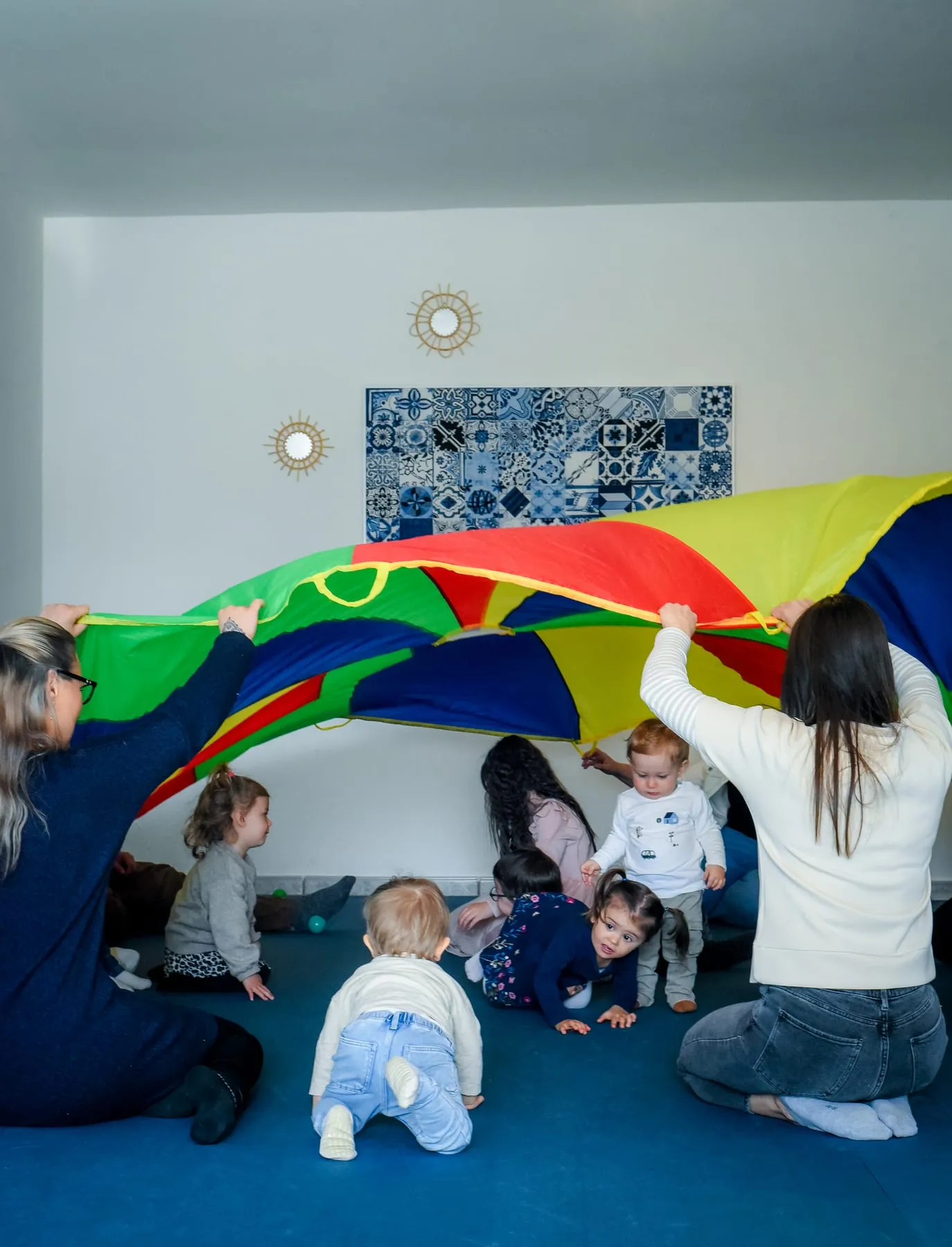 Zwei Frauen heben ein buntes Schwungtuch, darunter krabbeln, sitzen und stehen kleine Kinder