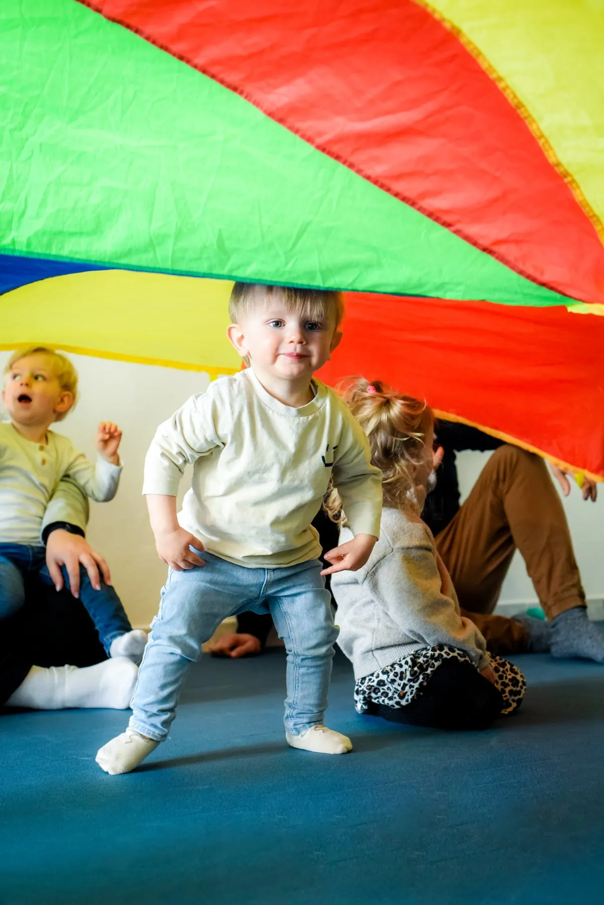 Unter einem bunten Schwungtuch steht ein Kleinkind mit gebeugten Knien, um es herum sitzen Kinder und Erwachsene auf dem Boden