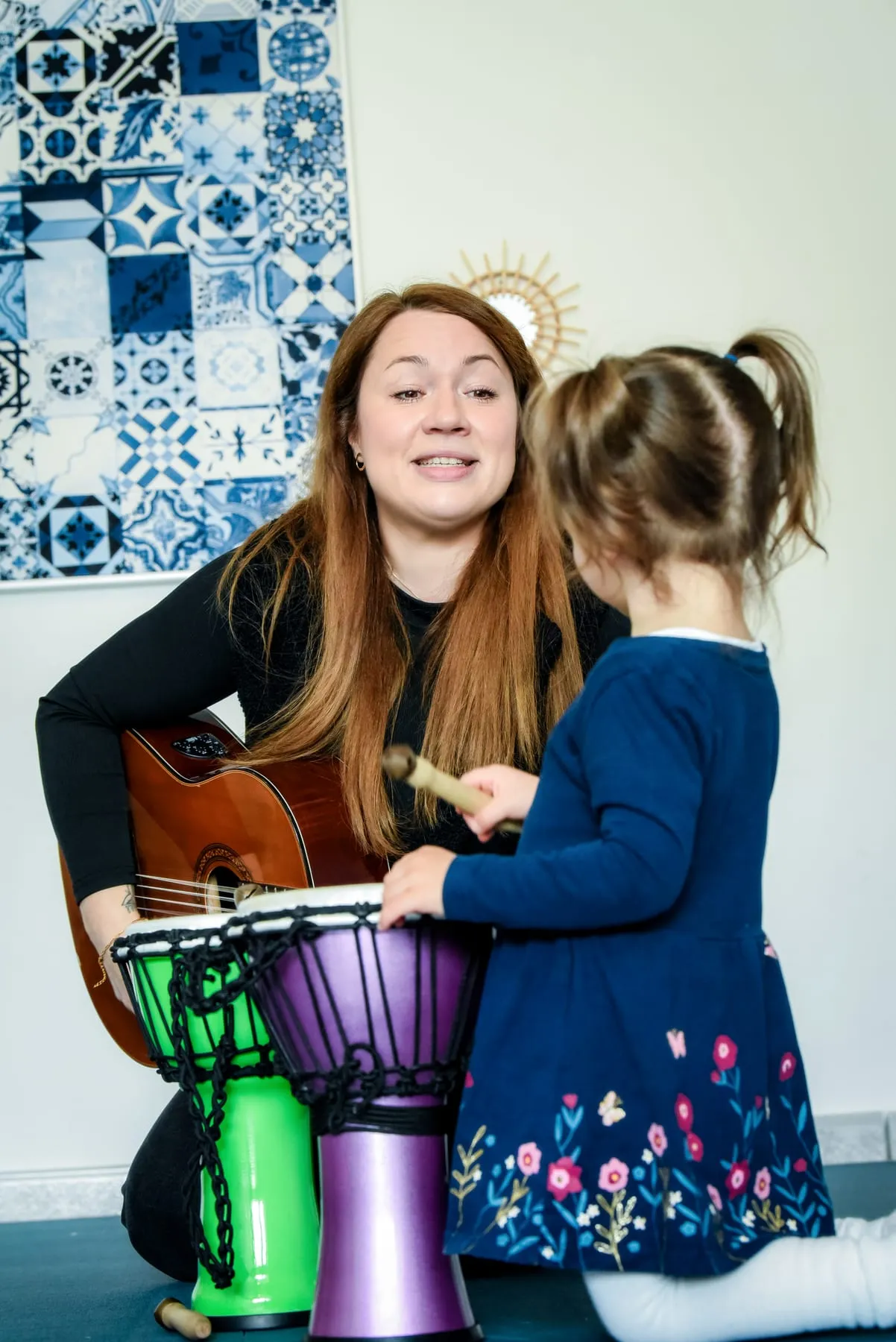 Eine Frau singt mit der Gitarre im Arm, vor ihr spielt ein kleines Mädchen mit einem Holzschlägel auf zwei bunten Djemben