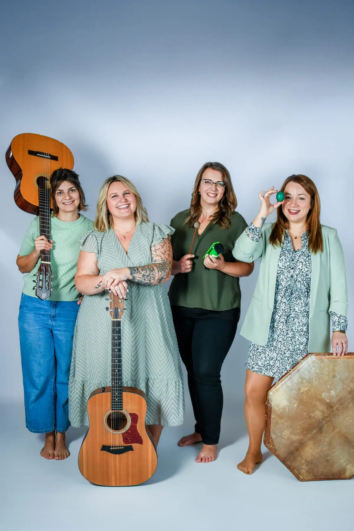 Vier Frauen des Teams mit zwei Gitarren, Rahmentrommel, Schlägel und Rassel-Ei vor grauem Hintergrund