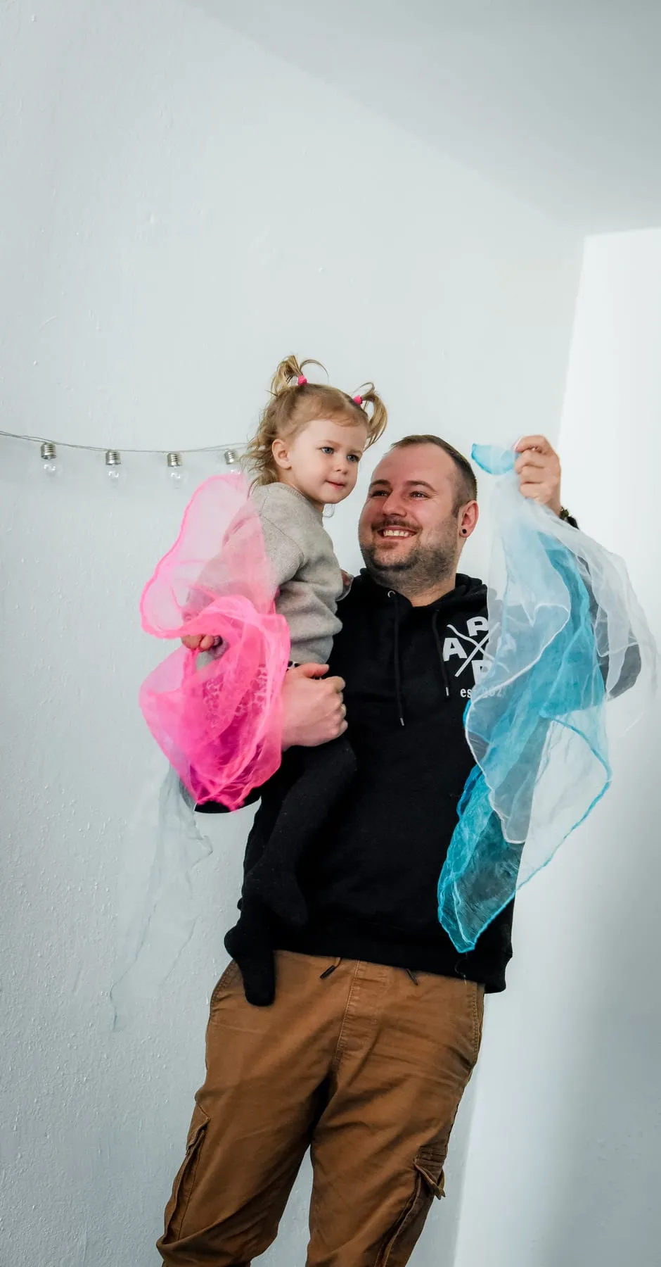 Ein Mann trägt ein kleines Mädchen auf dem Arm, beide halten bunte Tücher, er lacht
