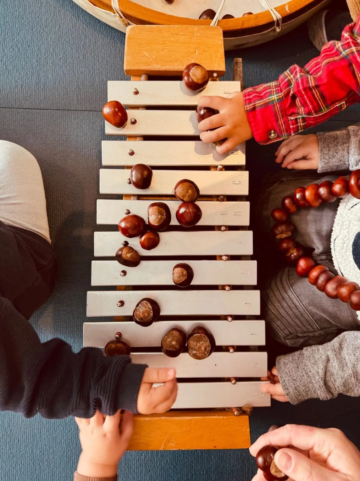 Von oben fotografiert: Auf einem Xylophon liegen Kastanien, Kinderhände greifen danach, rechts liegt eine Kette aus Kastanien, oben ist der Rand einer Rahmentrommel zu sehen